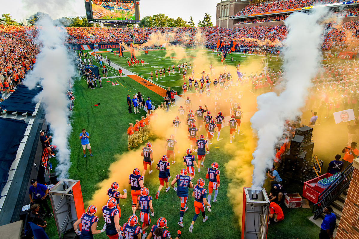 The Illini football team runs onto the field at Memorial Stadium from the tunnel surrounded by the band and orange smoke