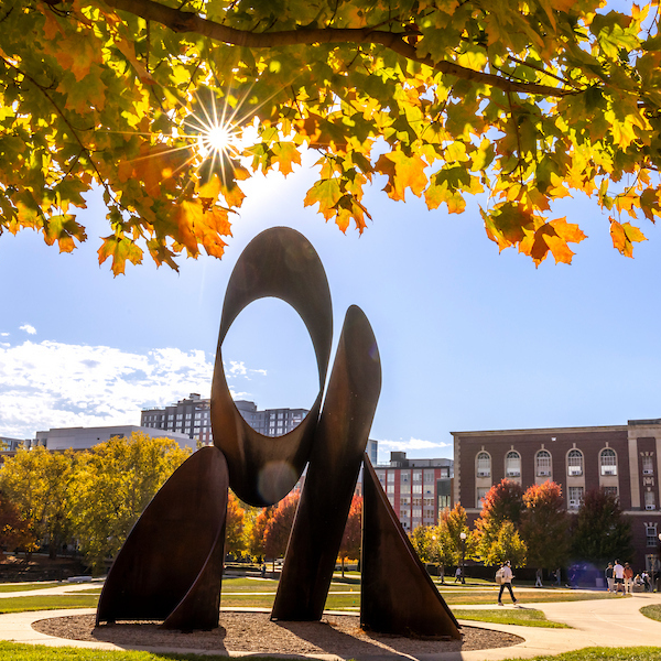 Image of a sculpture on the Bardeen Quad where there is fall foliage and the sun is shining