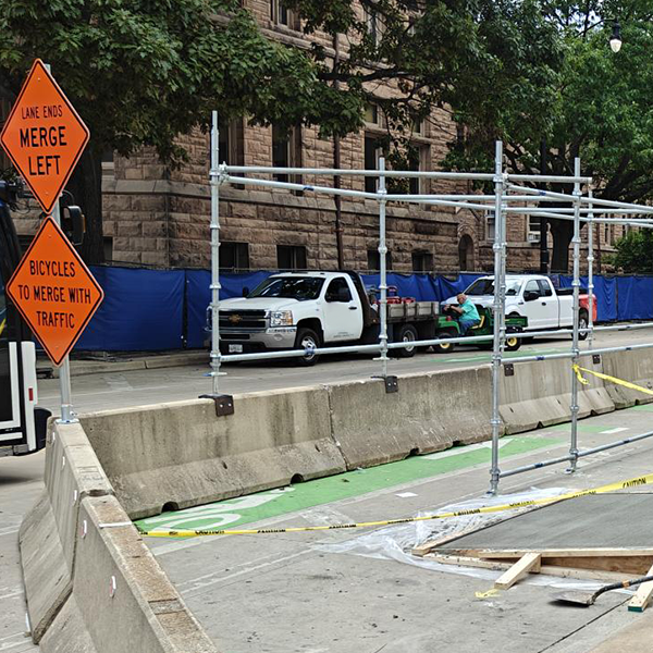 The Wright Street lane closure and covered walkway infrastructure as shown from Green Street. Two street signs indicate that a single lane must form and also that bicycles must merge left. Jersey barriers line the middle of Wright Street, forming the path for the covered walkway on the west side of the street.