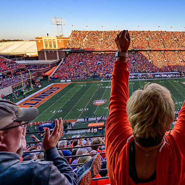 Illini fans cheering in Memorial Stadium at a football game - a view from behind an indvidual with both hands raised in the air