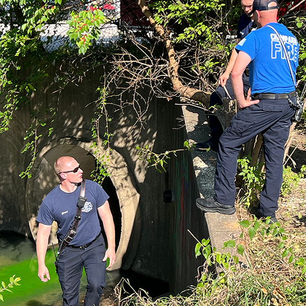 Several members of the Champaign Fire Department stand near a storm sewer inlet where the spill containment exercise is taking place. A green dye is in the water behind them.