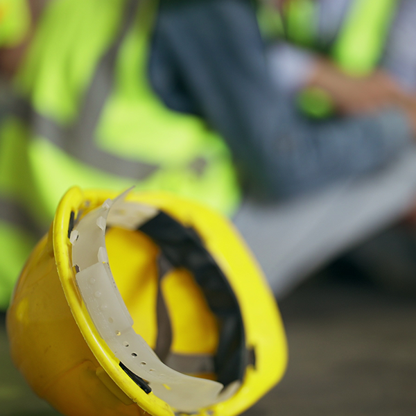 A yellow hard hat rests in the foreground as a worker with a safety vest sits in the background on a floor