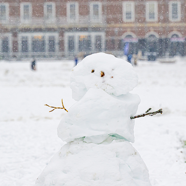 A snowman sits on the Main Quad with the Illini Union in the far background. The snowman's arms are sticks and other materials were used for eyes and a nose