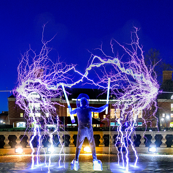 Terry Blake encounters bolts of electricity as he presents his Tesla Coil Concert event on the Bardeen Engineering Quad for the Engineering Open House. Technical note: Image consists of multiple exposures stacked into one image. The show features two giant Tesla coils, which generate bolts of electricity that fly through the air while simultaneously creating musical pitches. The coils, a heavily modified version of the device Nikola Tesla invented to wirelessly transport electricity, generate white-hot plasma. Protected in a suit of layered chain mail, Blake directs the bolts of electricity to fly across space using a variety of props, including neon tubes that glow like light sabers, flame throwers, and even a wooden 2x4 that quickly catches fire from the onslaught of electrical power.
