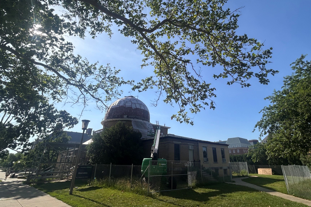 A construction crane is next to the outside of the Observatory. The site features a chain link fence and some scaffolding at the facility entrance.