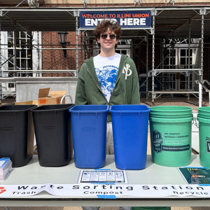 An EcoReps representative stands on the Illini Union's Anniversary Plaza in front of a table with various composting collection bins
