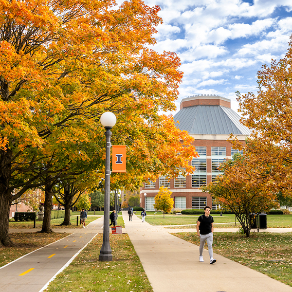 A student walks in front of the ACES Library, Information, and Alumni Center in the fall with trees changing colors.